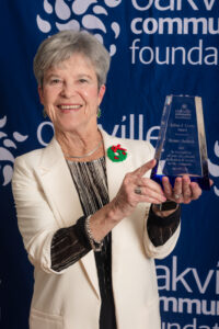 Bonnie Jackson poses with the Adrian Coote Volunteer Award in front of an Oakville Community Foundation banner.