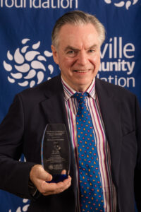 Michael Whitcombe poses with the Michael Whitcombe Award for Outstanding Volunteer in front of an Oakville Community Foundation banner.
